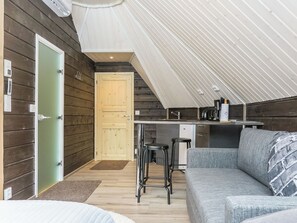A cozy interior with a wooden exterior, a kitchenette, a seating area with a sofa and bar stools, and a door leading to another room. - Arctic light hut by Interhome (Inari)