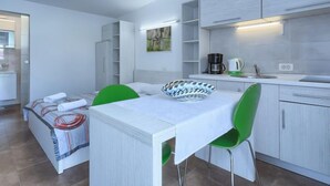 Ein stilvolles, helles Zimmer mit Esstisch, grünen Stühlen, einem weißen Sofa und einer Küchenzeile inklusive Kaffeemaschine und Wasserkocher.