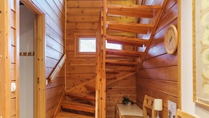 A wooden cabin interior with a staircase, a small table with chairs, and a clock on the wall.
