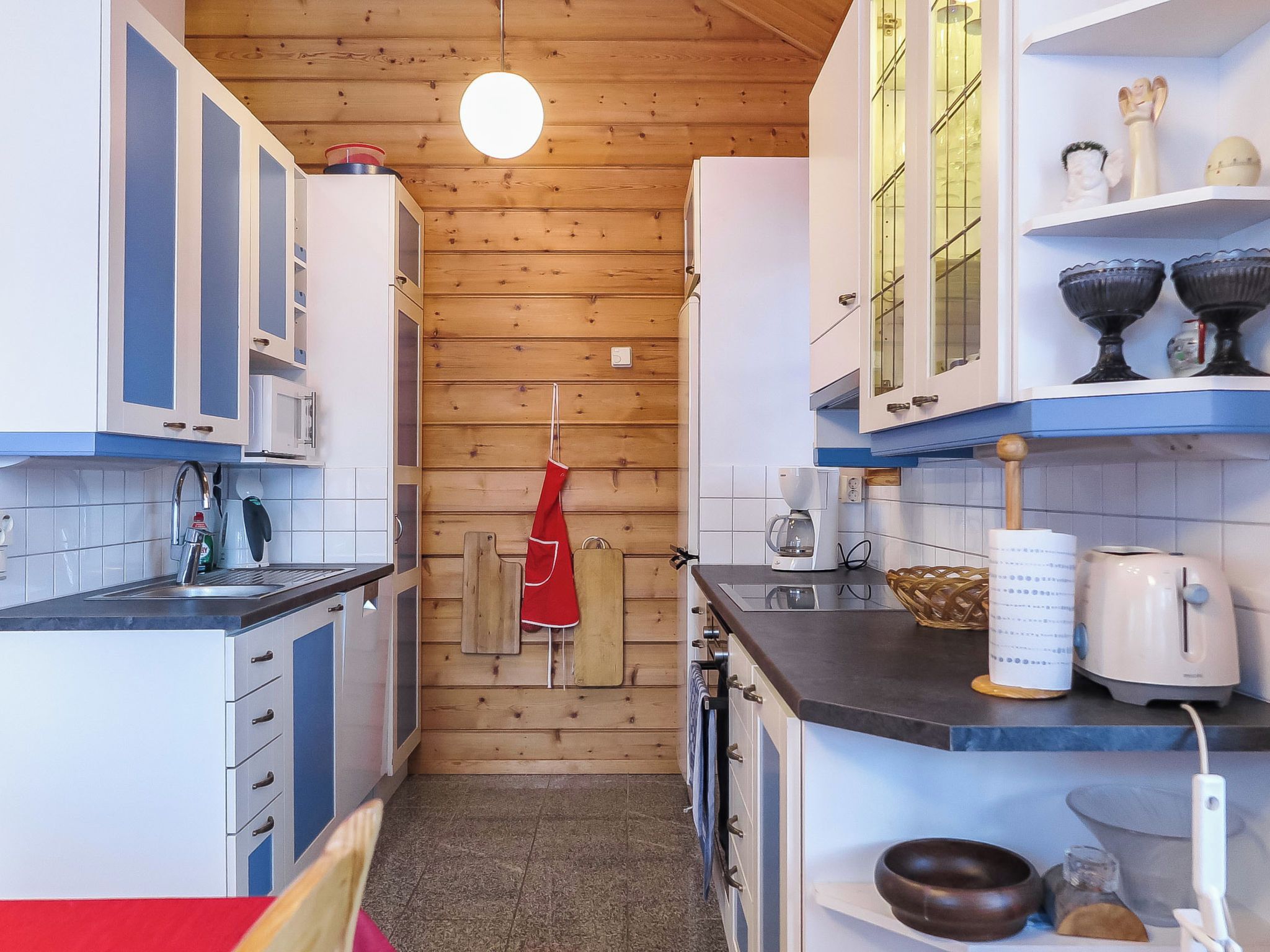 A compact kitchen with wooden walls, white cabinets, and a red apron hanging on the wall.