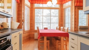 A cozy wooden cabin with a dining area featuring a red tablecloth, wooden chairs, and large windows with curtains.