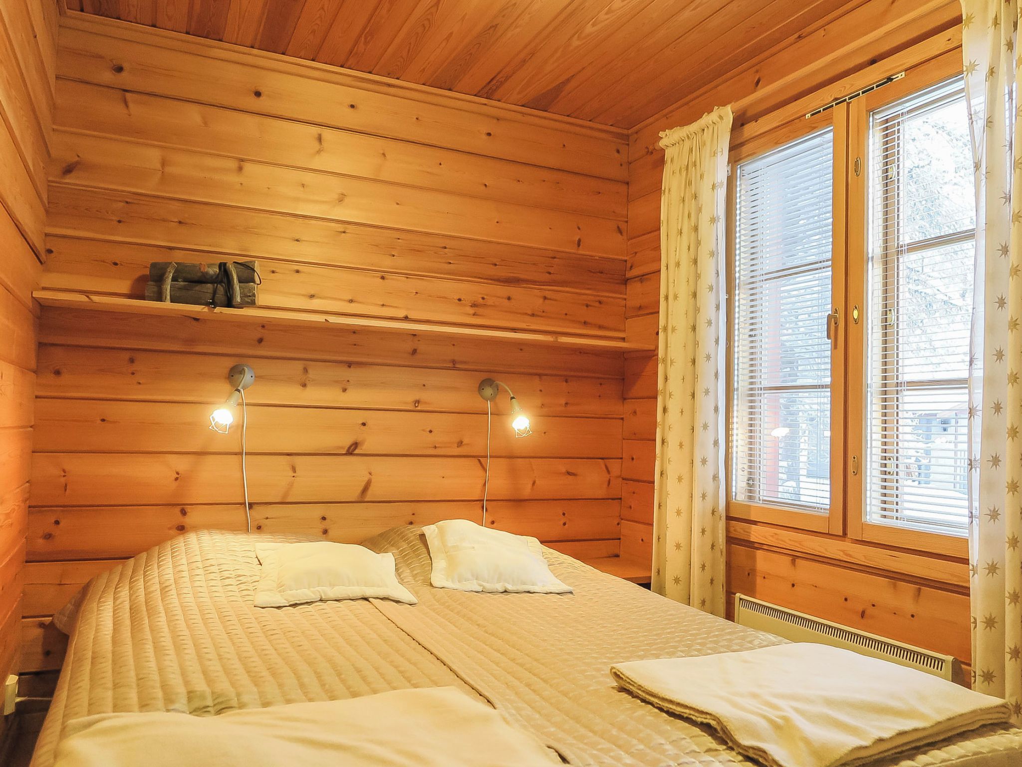 A wooden cabin room with a bed, two bedside lamps, a window with blinds, and a shelf with a bag.