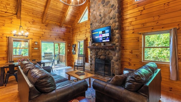 A log cabin interior with a stone fireplace, leather sofas, a television, and a dining area. - Bobcat Bluff-Hocking Hills State Park-Covered Hot Tub-Firepit-Pool Table-WiFi  (Logan)