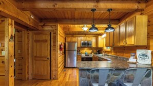 A wooden cabin interior with a stone island bar, wooden cabinets, and a stainless steel refrigerator. - Bobcat Bluff-Hocking Hills State Park-Covered Hot Tub-Firepit-Pool Table-WiFi  (Logan)