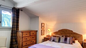 A bedroom with a wooden headboard, a bed with a white and purple bedspread, a wooden dresser, a nightstand with a lamp, and a window with a view of trees. - Chalet Le Renard Du Lac- close to the Chavanne piste and lake (Les Gets)