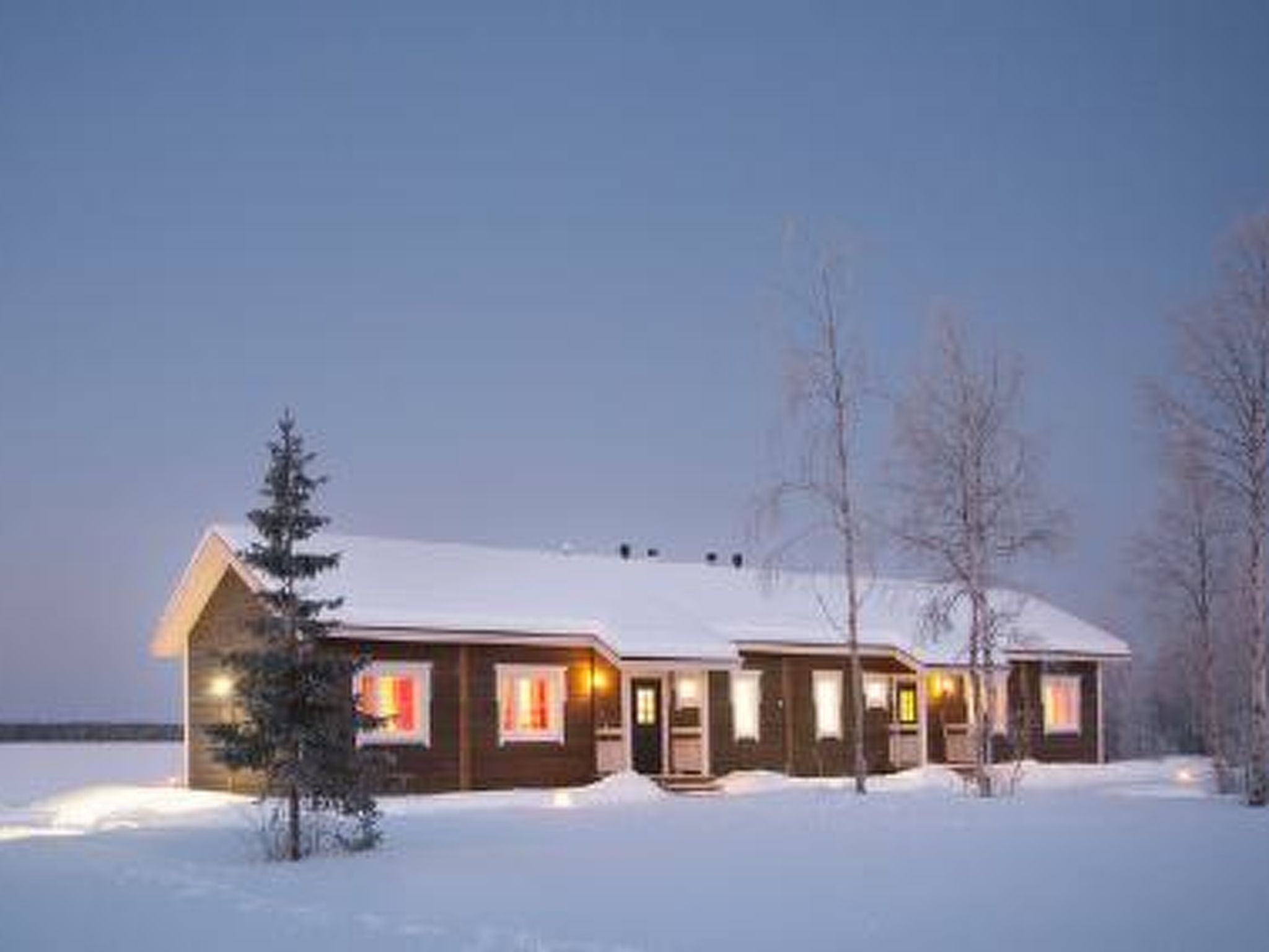 A snow-covered cabin with multiple windows and a porch, surrounded by bare trees and a snowy landscape.