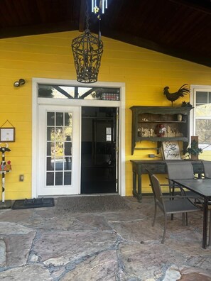 A yellow building with a white door, a rooster decoration, a sign that says 'Enjoy the Little Things,' and a stone patio area.