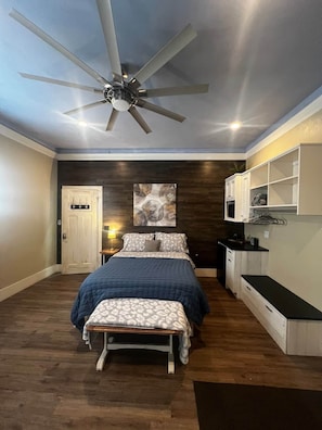 A bedroom with a bed, a ceiling fan, a nightstand, a wall-mounted shelf, and a door.