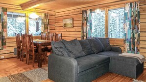 Interior acogedor de una cabaña con una mesa de comedor de madera, sillas, un sofá gris y un amplio ventanal con cortinas.