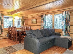 Interior de una cabaña de troncos con comedor, sofá y un amplio ventanal con cortinas.