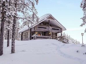 A wooden lodge with a steeply pitched roof, a front porch with a staircase, and a snow-covered landscape.