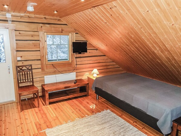 A cozy attic bedroom with a sloped ceiling, wooden walls, a bed, a TV, a chair, and a small table.