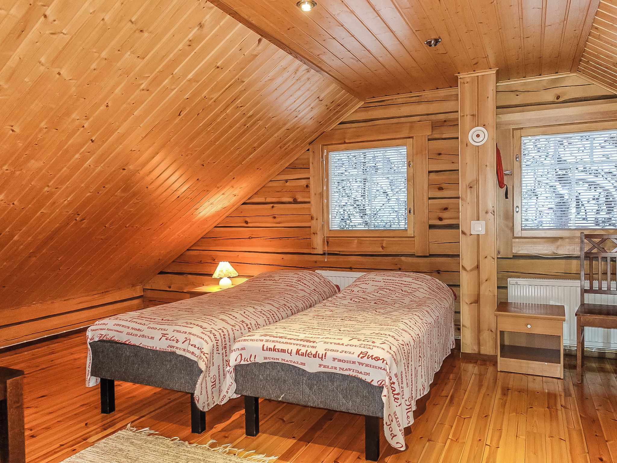 A cozy attic bedroom with a wooden sloped ceiling, a bed with a patterned blanket, a bedside lamp, and a window with a view of the outside.