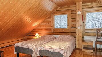 A cozy attic bedroom with a wooden sloped ceiling, a bed with a patterned blanket, a bedside lamp, and a window with a view of the outside.