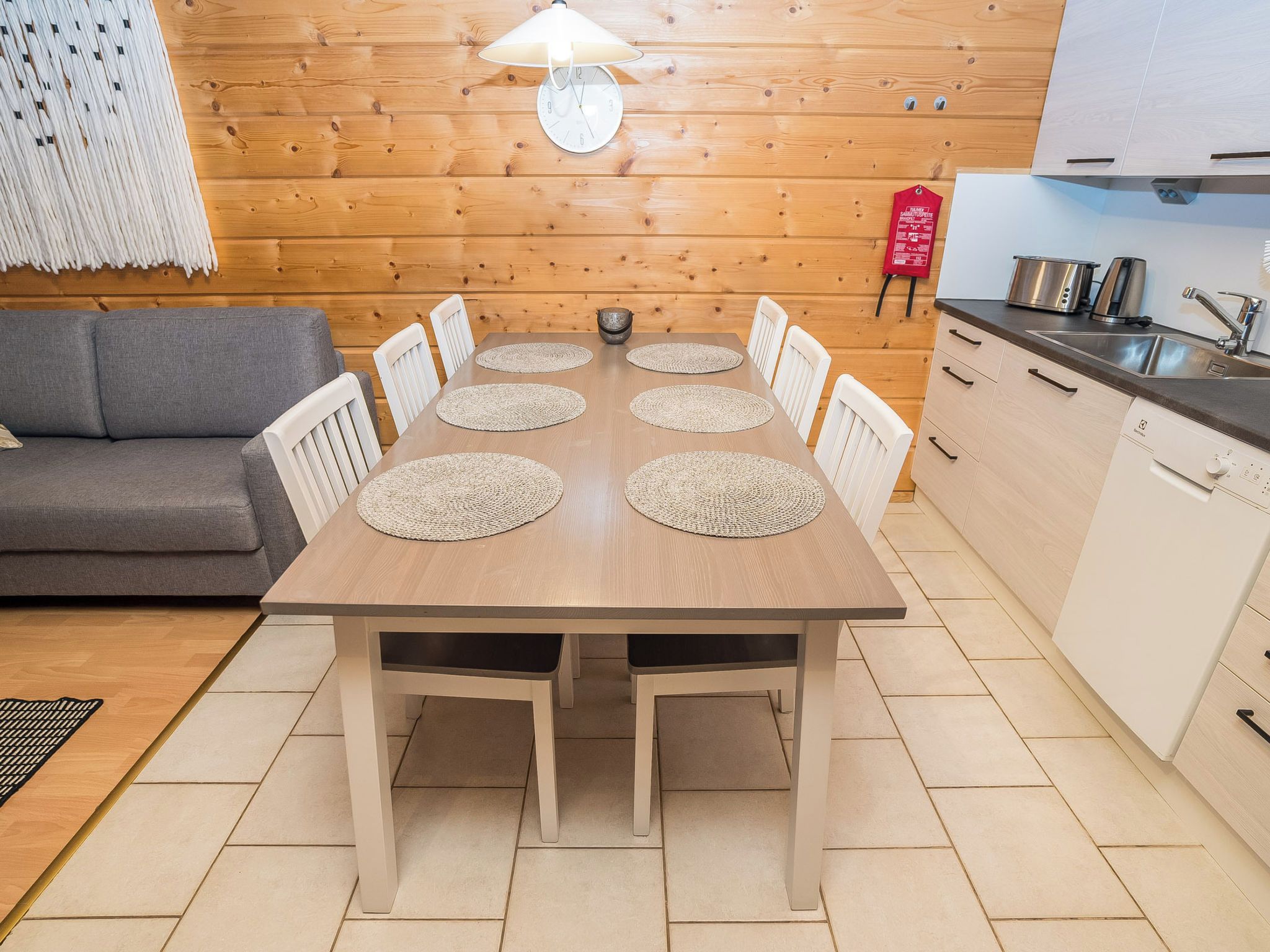 A dining table with six chairs, a kitchen area with a sink and stove, and a wooden wall with a clock.