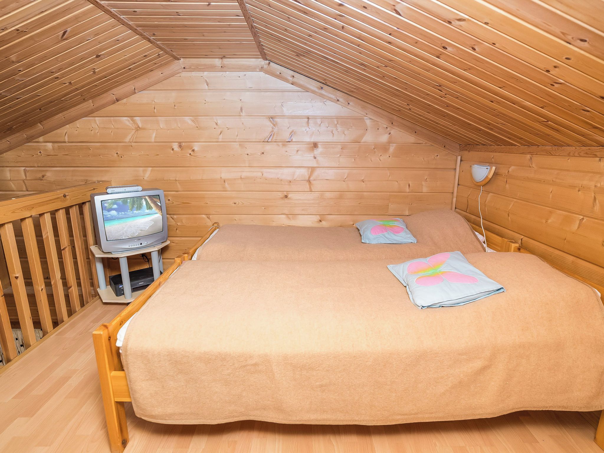 A small, cozy room with a wooden ceiling and walls, a single bed with a tan bedspread, a small table with a television, and a wall-mounted light fixture.