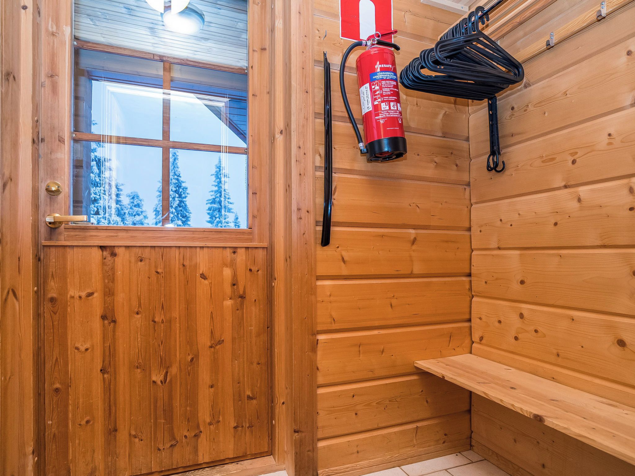 A wooden cabin interior with a door, a window, a fire extinguisher, and a bench.