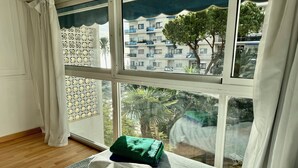 A room with a large window, a bed with a patterned bedspread, and a view of a residential area with trees and buildings. - Skol Apartments Marbella beachfront, central with views and WIFI - 2 bedrooms (Marbella)