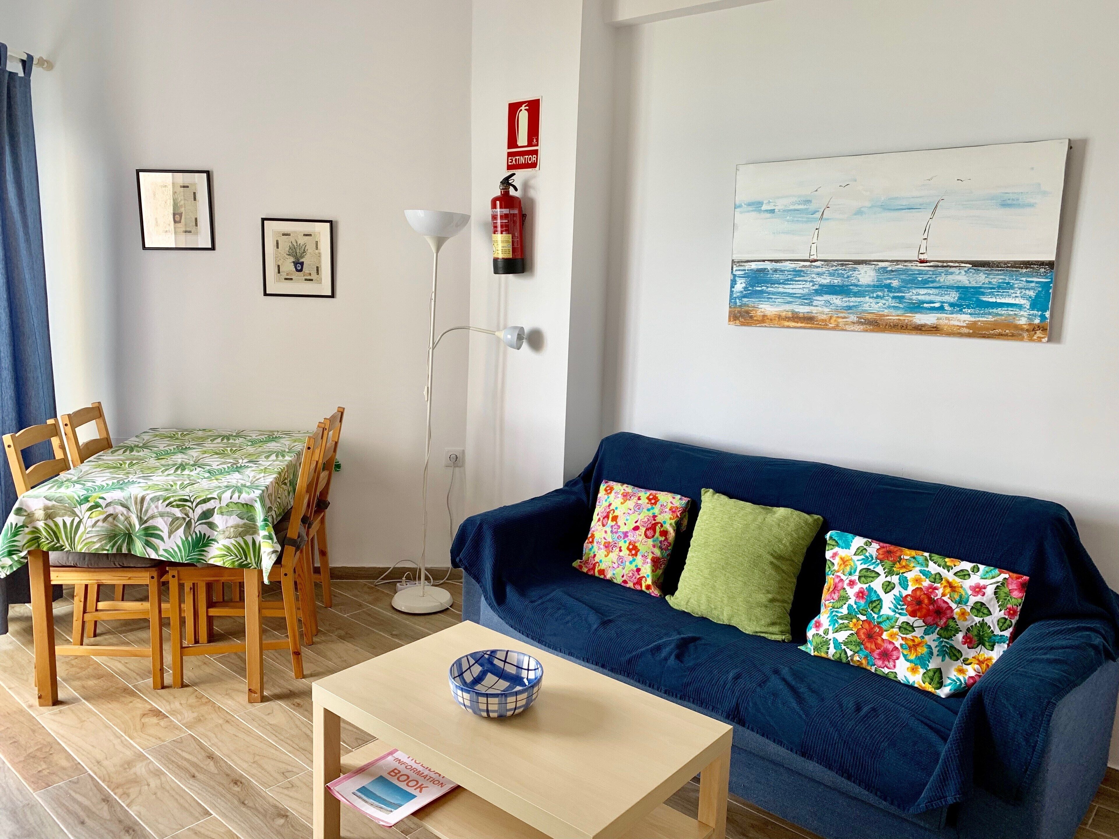 Ein Wohnzimmer mit einem blauen Sofa, einem Esstisch mit Stühlen, einer Stehlampe und einem Gemälde eines Segelboots an der Wand.