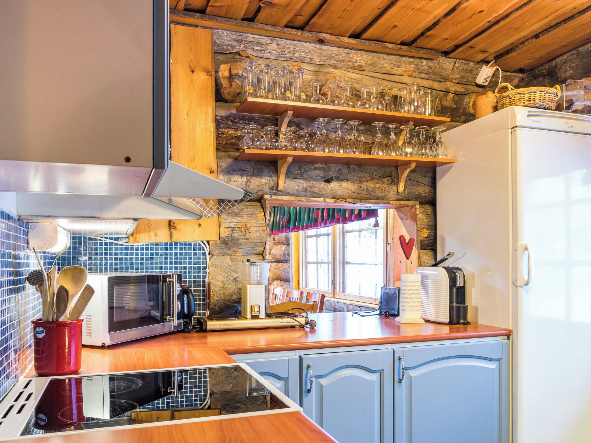 A kitchen with a microwave, toaster, and refrigerator. There is a wooden shelf with glassware above the counter.