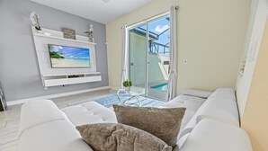 Un salon moderne avec un canapé blanc, un téléviseur à écran plat fixé à un mur blanc et une porte en verre donnant sur un espace piscine.