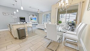 Une cuisine moderne dotée d’un four encastré, un coin repas avec une table en verre et des chaises blanches, et une grande fenêtre offrant une vue sur l’extérieur.
