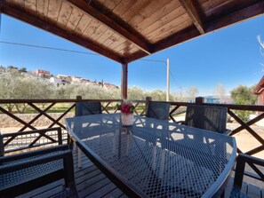 Un área de estar techada al aire libre con una mesa redonda de metal y sillas, una planta en maceta, y vista a edificios y árboles al fondo.