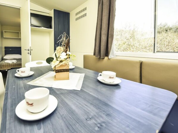 Habitación de hotel con una mesa de comedor preparada para dos comensales, una cama con cabecera, una ventana con cortinas y una planta pequeña sobre la mesa.