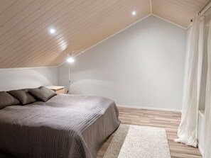 Ein Schlafzimmer mit schrägverdeckter Decke, einem Bett mit grauer Decke, einer Nachttischlampe und einem weißen Teppich.
