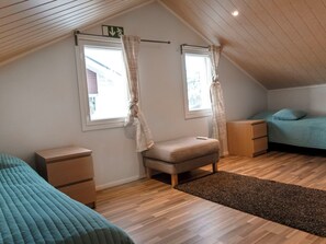 Una habitación con techo inclinado, dos ventanas con cortinas, una cama con colcha azul, un pequeño banco y una mesita de noche con lámpara.