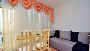 Un salon comprenant un canapé, deux fauteuils en bois, une table à manger et une fenêtre ornée de rideaux légers et de draperies orange.