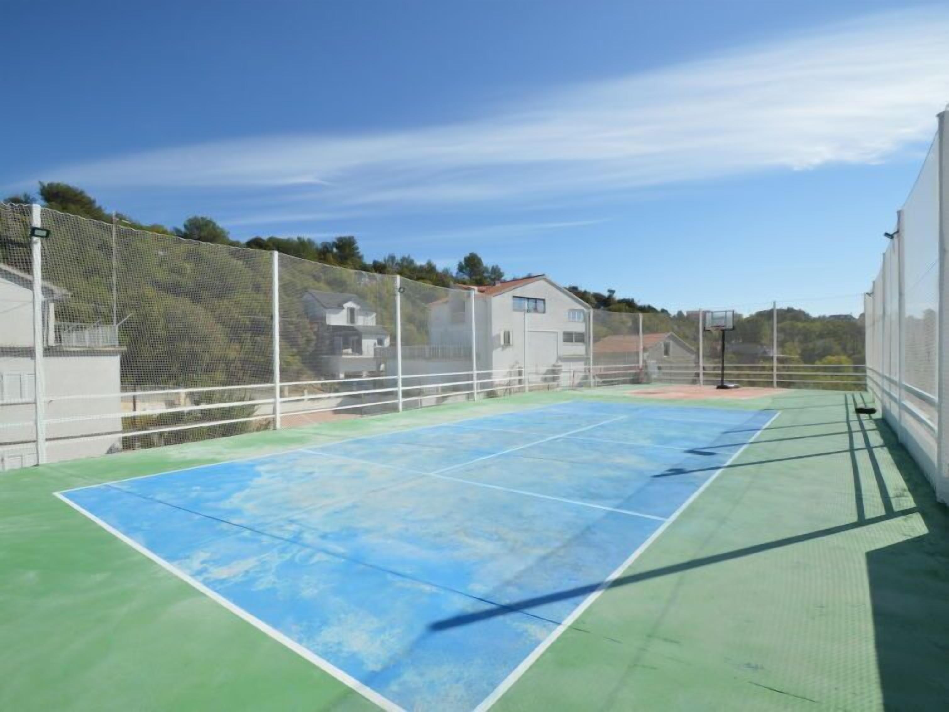 Un court de tennis en plein air, doté d’un filet, entouré d’une clôture et avec des maisons en arrière-plan.