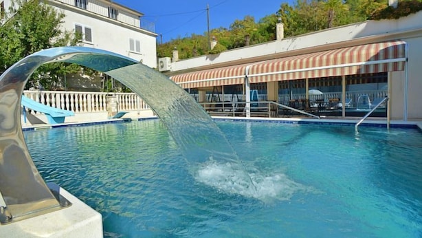 Une piscine agrémentée d’un élément décoratif aquatique, un espace salon abrité et un bâtiment en arrière-plan.