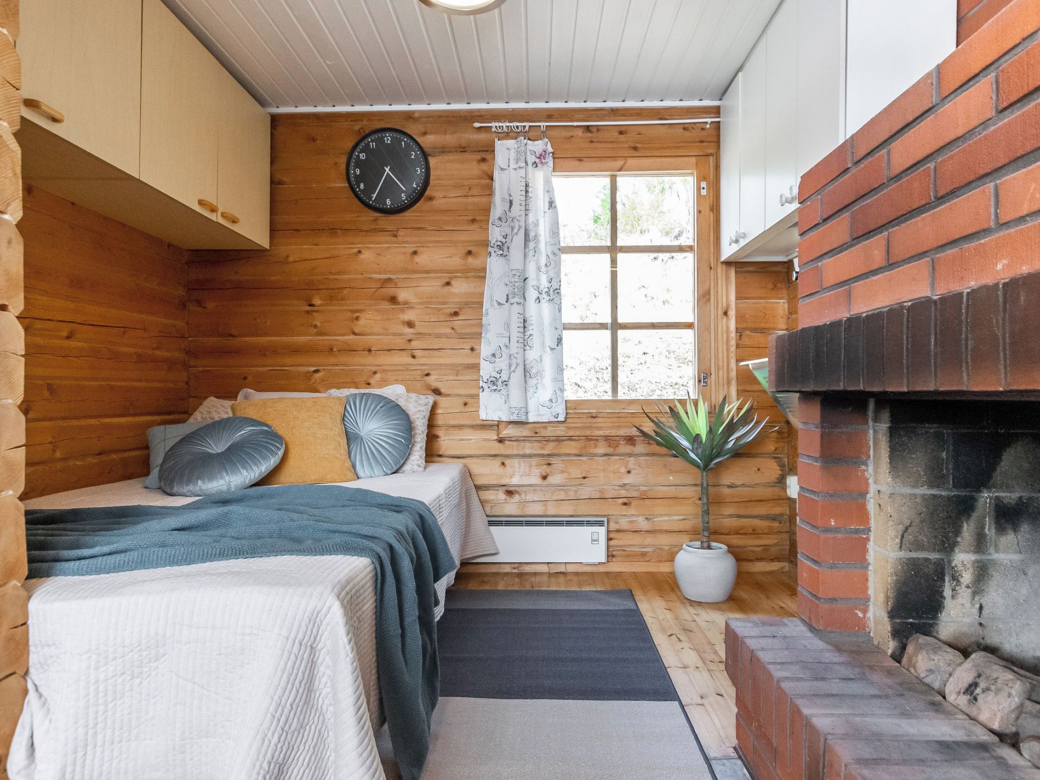A log cabin room with a bed, a clock, a window with curtains, a fireplace, and a potted plant.