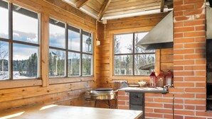 L’intérieur d’un chalet en bois comprend une grande table à manger, une cheminée en briques et de grandes fenêtres donnant sur un paysage enneigé.