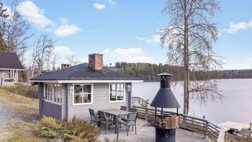 A lakeside cabin with a deck, outdoor seating, and a chimney.