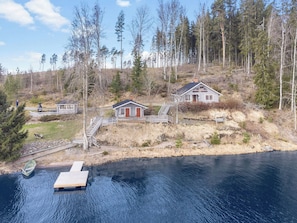 Un chalet au bord du lac, avec un quai, un petit bateau et une colline boisée.