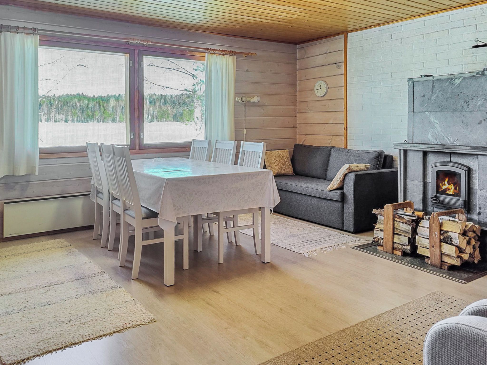 Ein gemütliches Blockhaus-Interieur mit Esstisch, Stühlen, einer Couch, einem Kamin und Blick ins Freie.
