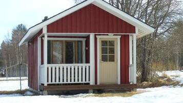 A small, single-story red cabin with a white porch and a door, surrounded by snow.