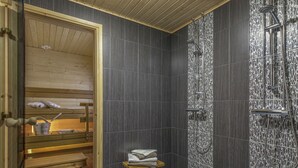 Un cuarto de sauna con un banco de madera, un taburete pequeño con toallas y una zona de ducha con un detalle decorativo en la pared.