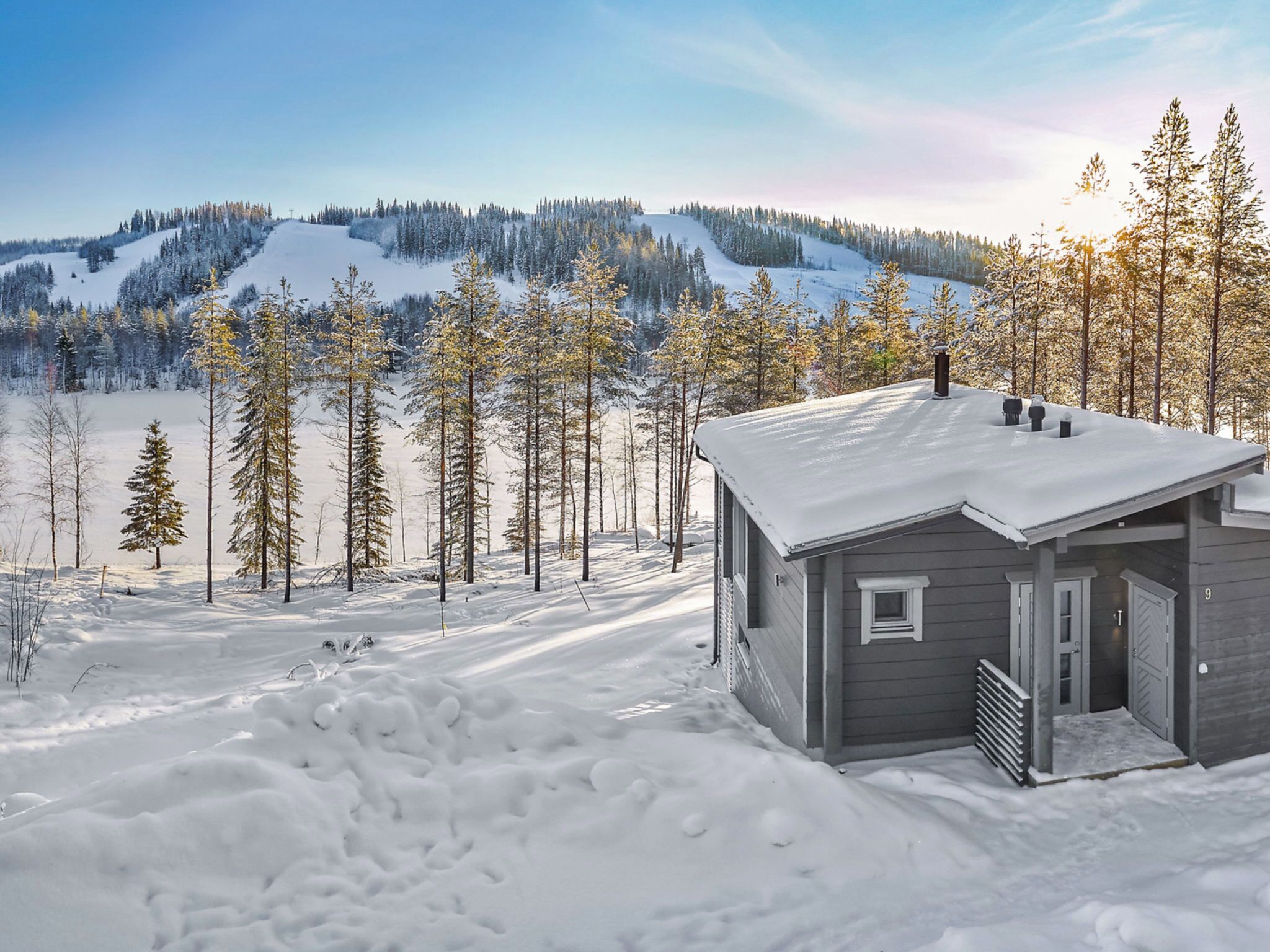 A cabin with a snow-covered roof and a small window, surrounded by a snowy landscape and trees.