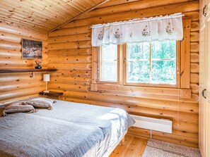 Puisesta materiaalista rakennettu mökkimäinen huone, jossa on sänky, ikkunassa kukkakuviollinen verho, patteri ja seinällä taulu.