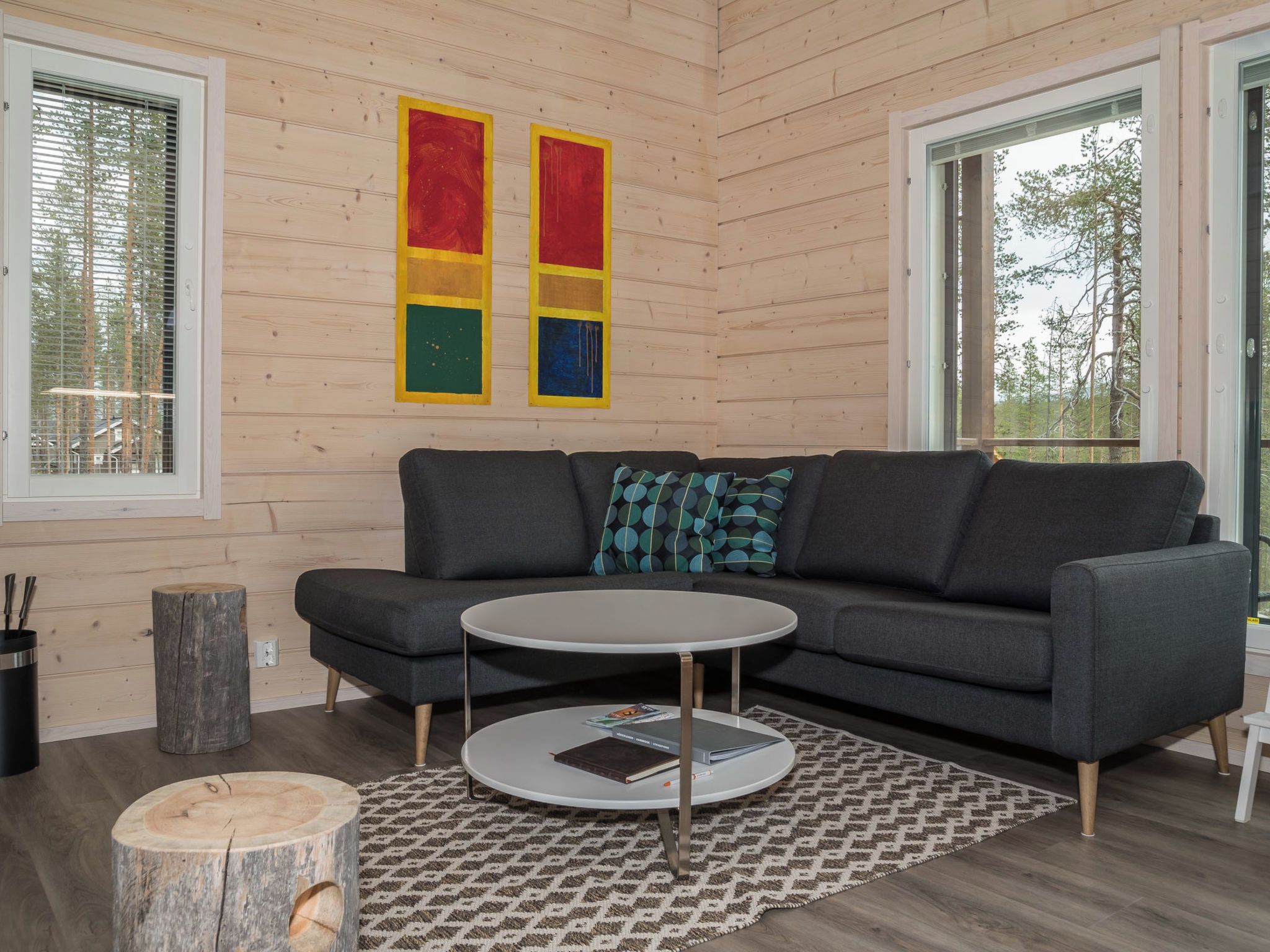 A modern living room with a grey sofa, a round coffee table, and a patterned rug. There are two windows with blinds, a wooden wall, and a painting above the sofa.