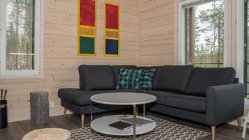 A modern living room with a grey sofa, a round coffee table, and a patterned rug. There are two windows with blinds, a wooden wall, and a painting above the sofa.