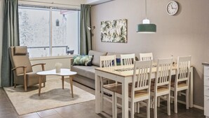 Een moderne woonkamer met een eethoek, een bank, een salontafel en een eettafel met stoelen. Er is een raam met gordijnen, een klok aan de muur en een hanglamp.