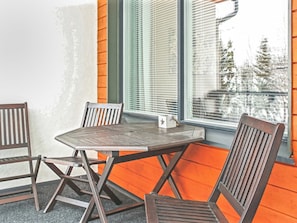 Un coin salon avec une table en métal et des chaises en bois, une fenêtre avec des stores et une vue sur les arbres à l’extérieur.