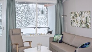 Een moderne woonkamer met een grijze bank, een bruine fauteuil en een witte salontafel. Er is een raam met uitzicht op met sneeuw bedekte bomen.