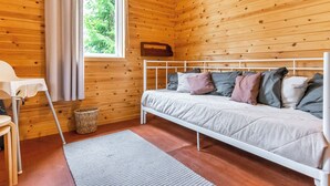 Habitació d'una cabana de fusta amb un llit, una finestra amb cortines, una taula petita i una cistella.