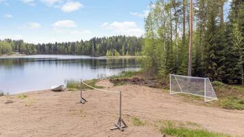 A sandy beach with a goalpost, a calm lake, and a forest in the background.