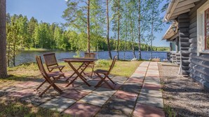 En stuga vid sjön med en uteplats med ett träbord och stolar, omgiven av träd och med utsikt över sjön.
